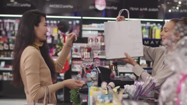 Średni widok z boku strzał z powolnym tempie atrakcyjnej azjatyckiej kobiety stojącej w kasjerce w sklepie makijażu, gdy asystentka na zakupach daje jej torbę z zakupionymi kosmetykami uśmiechając się do siebie - Materiał filmowy, wideo