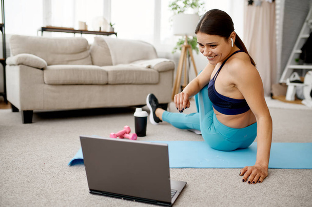 A mosolygó lány otthon ül a padlón, online edzést tart a laptopnál. Női személy sportruházat, internetes sport edzés, belső tér a háttérben - Fotó, kép