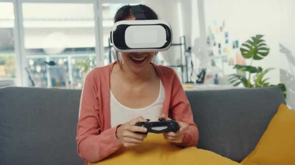 アジアの女性は、自宅のリビングルームでソファの上で仮想現実のプレイジョイスティックゲームのヘッドセットメガネを着用してください。ホームcovid隔離に滞在し、現実を再想像し、自宅でVR 、将来の概念のVR技術. - 映像、動画