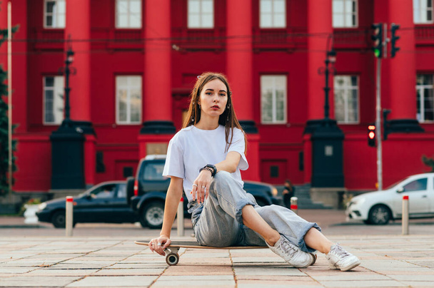 Relajada hermosa patinadora joven está sentada en su monopatín, posando para una foto y mirando a la cámara delante de un edificio rojo. - Foto, imagen