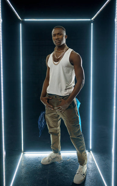 Seriöser Rapper in Goldschmuck posiert vor beleuchtetem Kubus, dunklem Hintergrund. Hip-Hop-Performer, Rap-Sänger, Breakdance-Performer, Unterhaltungskünstler, Breakdancer - Foto, Bild