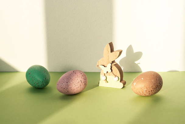 Houten speelgoedkonijntje en beschilderde eieren op een groene tafel tegen een witte muur met schaduwen. Pasen concept. - Foto, afbeelding