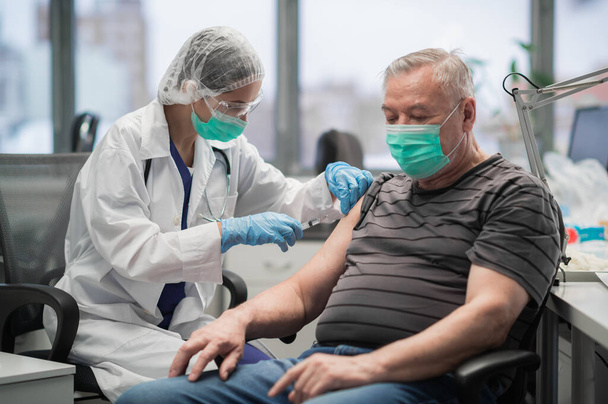 Een jonge verpleegster met een masker geeft een coronavirusvaccinatie aan een oudere man in een kliniek - Foto, afbeelding