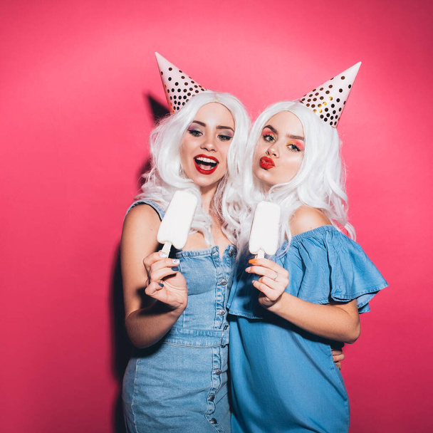 Two cute girls in white color wigs and denim dresses on a neutral pink background. They are licking ice creams happily. Smiling lifestyle portrait with harsh fashion lighting - Photo, Image
