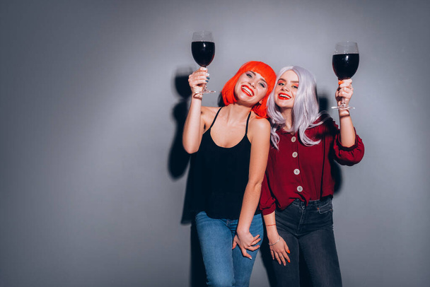 Dos lindas chicas borrachas en pelucas rojas y grises vestidas con jeans y blusas sobre un fondo gris neutro. Están bebiendo vino felizmente. Retrato de estilo de vida sonriente con iluminación de moda dura - Foto, Imagen