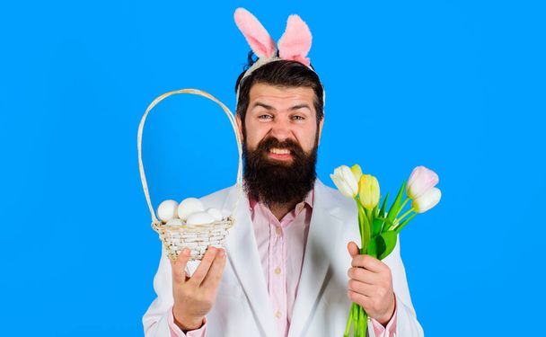 Vacanze di Pasqua. Uomo barbuto con orecchie di coniglio con uova di cesto e bouquet di tulipani. Un coniglietto. Caccia alle uova. - Foto, immagini