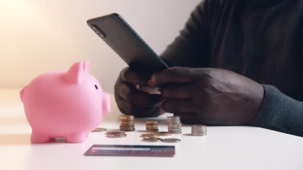 Een man gebruikt telefoon om zijn bankrekening te controleren. Piggy bank credit card en munten op de tafel. Investeringen via mobiele app - Video