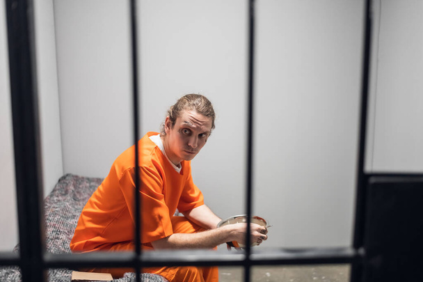 Ένας κρατούμενος υψηλής ασφάλειας με κόκκινη στολή φυλακής τρώει από ένα σιδερένιο μπολ στο κελί του.. - Φωτογραφία, εικόνα