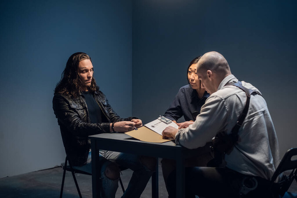 Un duro detective de policía interroga a un sospechoso de crimen con una cara tatuada en una sala de interrogatorios. - Foto, imagen