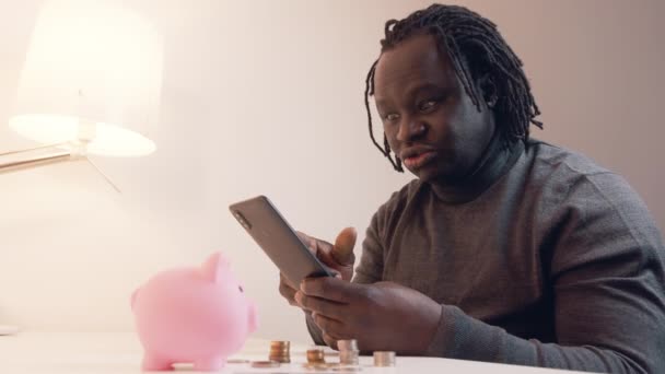 銀行口座を確認するのに電話を使った男。テーブルの上の貯金箱のクレジットカードとコイン。モバイルアプリを使った投資 - 映像、動画