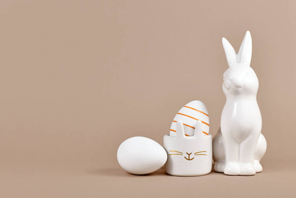 Golden striped Easter egg in bunny egg cup next to rabbit figure and white egg on side of beige background with empty copy space - Photo, Image