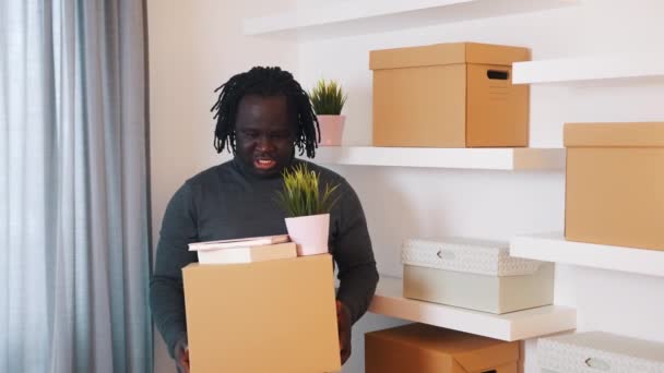 Young african american man relocating in to the new appartment. Holding carboard boxes and plant - Footage, Video