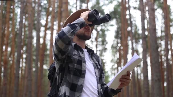 Een man met een hoed houdt een kaart vast en kijkt door een verrekijker terwijl hij door het bos wandelt. Oriëntatieconcept, reizen. - Video
