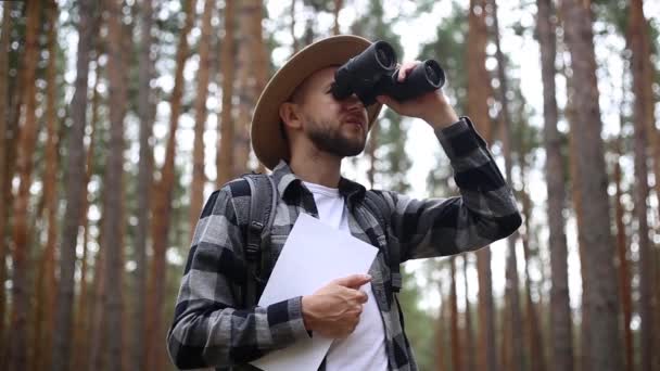 帽子の男はマップを保持し、森の中でハイキング中に双眼鏡を見ています。オリエンテーションコンセプト、旅行. - 映像、動画
