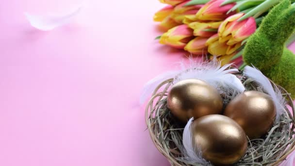 Huevos de Pascua Color dorado en cesta con tulipanes primaverales, plumas blancas sobre fondo rosa pastel en feliz decoración de Pascua. Decoración festiva - Metraje, vídeo