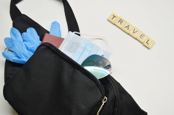 Černá taška s pasem, lékařskou maskou, modrými gumovými rukavicemi a slunečními brýlemi na bílém pozadí. Cestovní slovo z dřevěných kostek - Fotografie, Obrázek