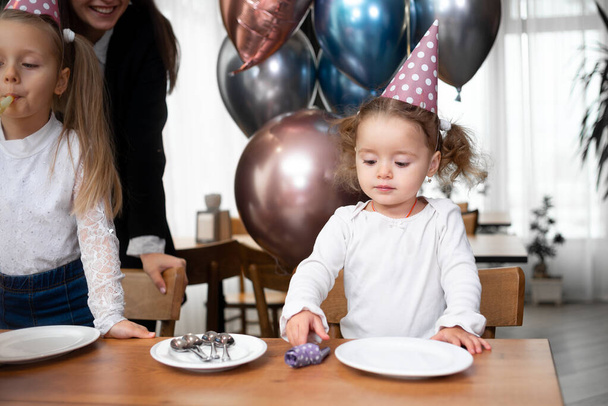 Ευτυχισμένη οικογένεια γιορτάζει γενέθλια ή επέτειο. Κοριτσάκι με εορταστικό καπέλο. Τούρτα γενεθλίων και μπαλόνια. - Φωτογραφία, εικόνα