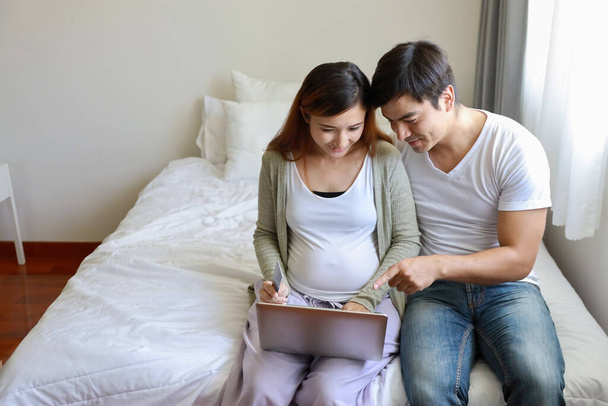 İnternetten alışveriş yapmak için kredi kartı tutan genç, hamile Asyalı bir kadın. Kafkasyalı kocasıyla yatak odasında kucaklaşan, gülen yüzlü bir bilgisayar kullanıyor.  - Fotoğraf, Görsel