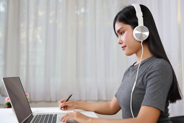 Mladá chytrá a aktivní asijská studentka sedí u stolu se sluchátky, používá počítač a bere na vědomí pro studium on-line se šťastnou tváří (nový normální koncept) - Fotografie, Obrázek