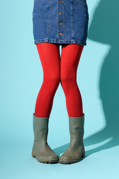 Αστεία πόδια μιας νεαρής γυναίκας με denim φούστα, κόκκινο κολάν και πράσινες λαστιχένιες μπότες που ποζάρουν με χτυπημένα γόνατα και πόδια περιστεριού πάνω από ένα μπλε φόντο με copyspace - Φωτογραφία, εικόνα