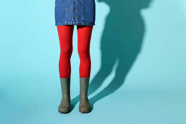 Jovem mulher em botas de borracha verde ou gumboots vestindo uma saia jeans curto e meia-calça vermelha ou colante posando sobre um fundo azul com copyspace e sombra em uma visão inferior do corpo - Foto, Imagem