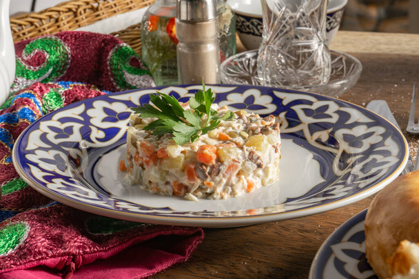 Klasik Olivier salatası, mayonez, sebze ve sosis seramik tabakta maydanozla süslenmiş geleneksel Özbek süslemeleri ile birlikte çatal ve bıçaklı çatal ve bıçakla birlikte ahşap bir masa üzerinde.. - Fotoğraf, Görsel