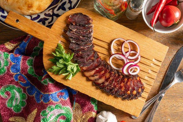 Rebanadas de embutido casero de carne de caballo seca con cilantro y cebolla roja, cortadas en rodajas y servidas en una tabla de picar de madera - Foto, imagen