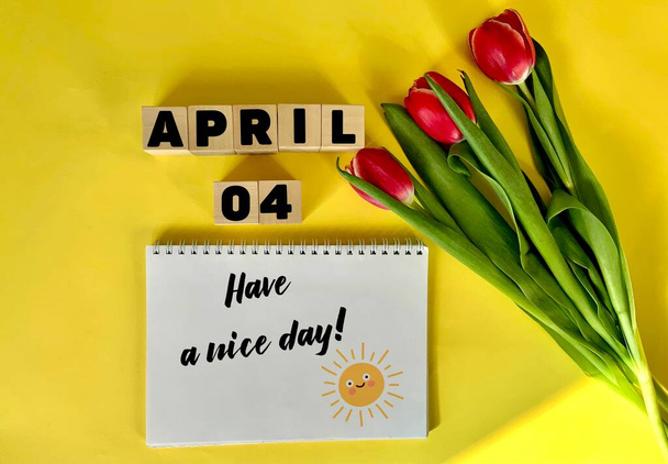 4 апреля на деревянных кубиках на желтом фоне. Рядом с букетом тюльпанов и блокнотом с надписью хорошего дня. Календарь на апрель . - Фото, изображение