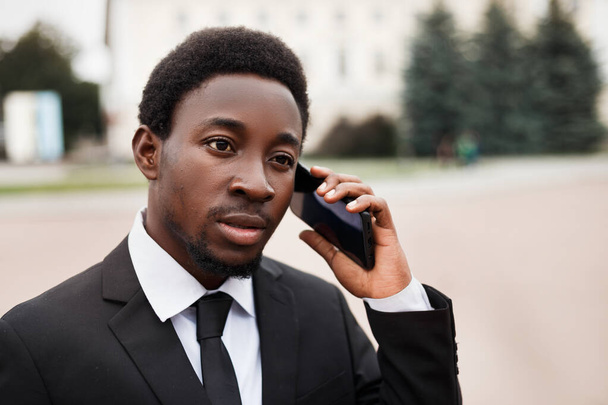 Concentration sélective du jeune homme d'affaires afro-américain parlant au téléphone. Homme au visage sérieux utilisant un gadget dehors. Concept d'entreprise avec espace de copie. - Photo, image