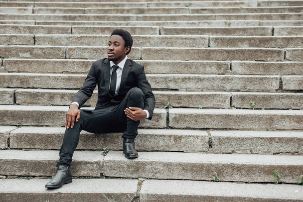 Jeune homme d'affaires afro-américain porter costume noir et assis sur les escaliers, regardant loin. - Photo, image