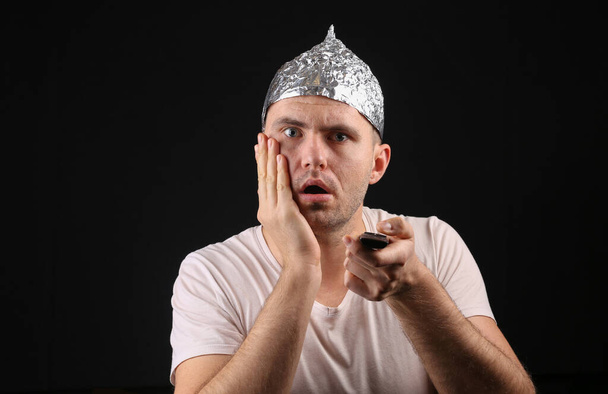 Hullu mies alumiinifolio hattu käyttäen tv kaukosäädin mustalla pohjalla - Valokuva, kuva
