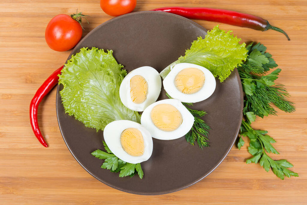 Metades de ovos fervidos com folhas de alface, endro e salsa galhos em prato marrom entre de verduras em uma superfície de madeira, visão superior - Foto, Imagem