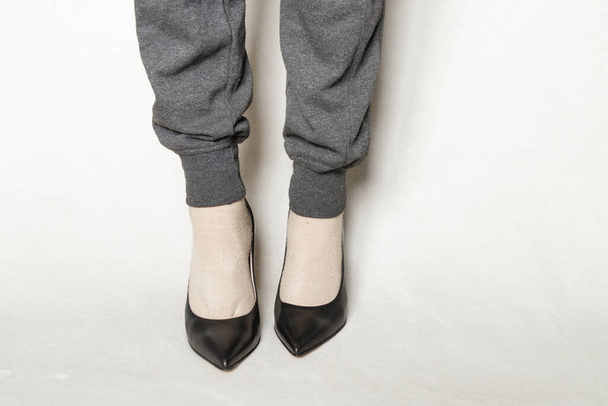una giovane ragazza in pantaloni della tuta grigia e scarpe con tacco alto punta nera si erge su un tappeto bianco a casa - Foto, immagini