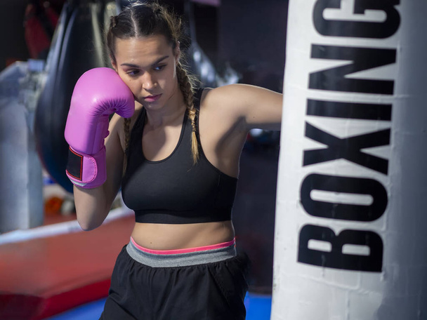 Boxer nő üt egy bokszzsák rózsaszín kesztyűvel a tornateremben edzés és edzés közben. Magabiztos nő francia fonatokkal, bokszolást és önvédelmet gyakorol. Lány teljesítmény és fitness koncepció - Fotó, kép