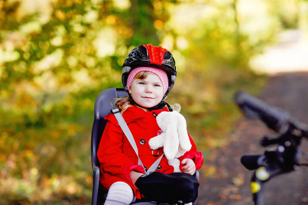 Malá batolátko dívka s bezpečnostní helmou na hlavě sedí na kole sedadla své matky nebo otce kolo. Bezpečná a koncepce ochrany dětí. Rodinný výlet na podzim. Dítě drží měkkou hračku - Fotografie, Obrázek