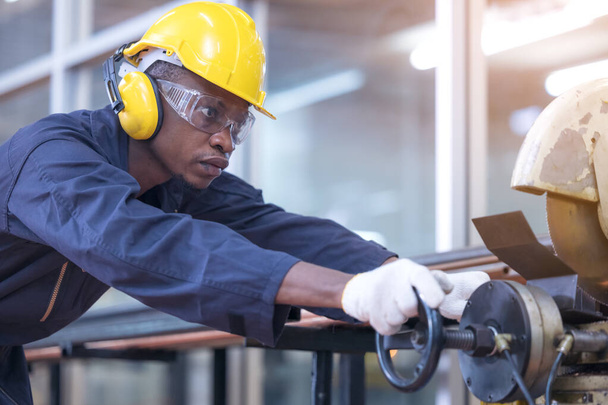 Чёрный мужчина, африканский рабочий, носит звуконепроницаемые наушники и жёлтый шлем, работающий на заводе по резке железа.. - Фото, изображение
