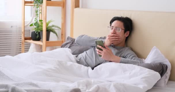 Hombre bostezando y estirándose en la cama mientras usa el teléfono móvil - Imágenes, Vídeo