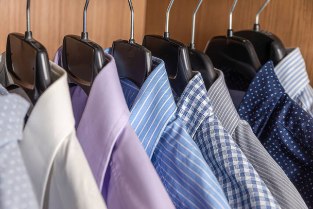 Hemden in verschiedenen Farben, nebeneinander auf Kleiderbügeln in einem holzfarbenen Kleiderschrank aufgehängt - Foto, Bild