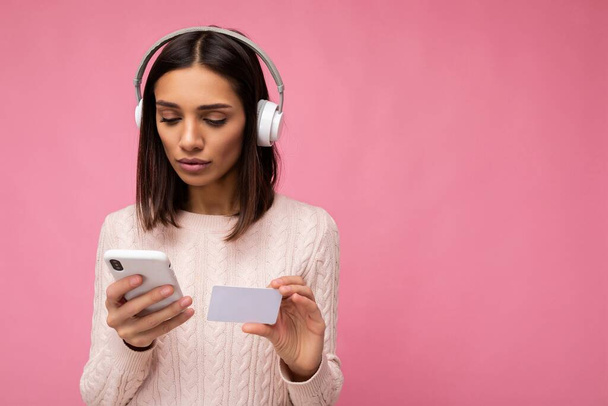 Kuva kaunis nuori brunette nainen yllään vaaleanpunainen rento villapaita eristetty vaaleanpunainen taustaseinä yllään valkoinen bluetooth langattomat kuulokkeet ja musiikin kuuntelu ja matkapuhelimen tekeminen - Valokuva, kuva