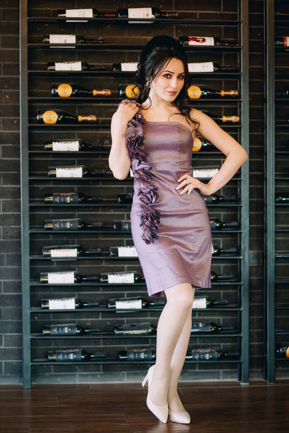 Πορτρέτο της πολυτέλειας κορίτσι με μωβ φόρεμα στέκεται μπροστά από τα ράφια με κρασί και σαμπάνια μπουκάλια στο εστιατόριο - Φωτογραφία, εικόνα