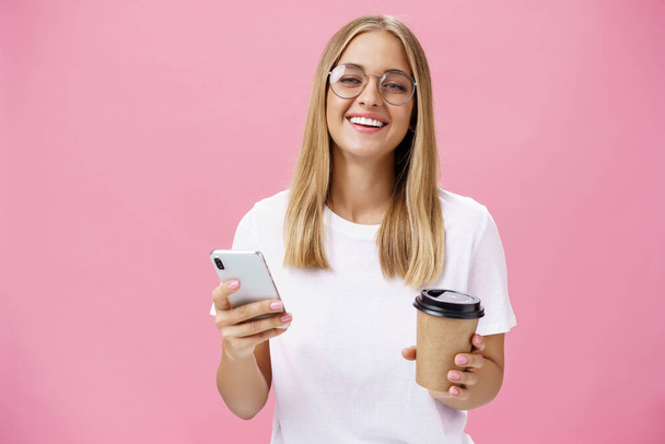 Busy freelancer sipping coffee and working via smartphone talking to clients while sitting in cafe or coworking place, laughing joyfully at camera with broad smile holding paper cup and cellphone - Foto, imagen