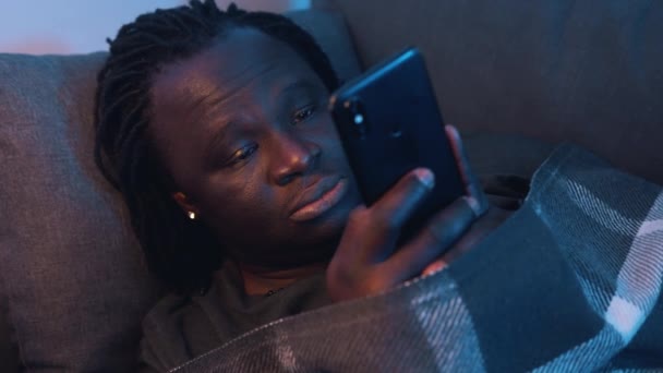 Unatkozó fekete férfi pihentető a kanapén csomagolva a takaró és az okostelefon használata - Felvétel, videó