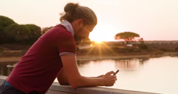 Kaukázusi fickó piros pólóban sms-ezik naplemente égbolttal a háttérben - Felvétel, videó