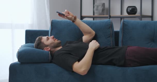 mladý muž komunikuje prostřednictvím videohovoru v mobilním telefonu, leží na gauči doma, dálkové komunikace a internetové technologie - Záběry, video