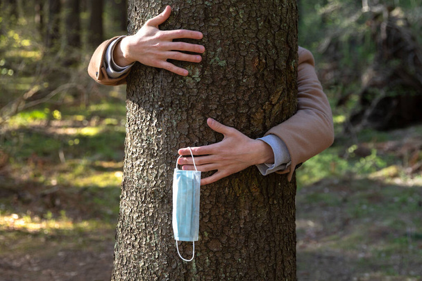 Close-up van de handen omhelzing van een boom en doet de medische bescherming masker af, houdt het op haar vingers, geniet van het leven, schone frisse lucht in het bos na Covid-19 pandemie, zelf-isolering.Quarantaine is voorbij - Foto, afbeelding