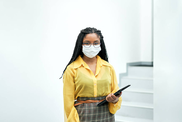Retrato de una mujer de negocios mirando a la cámara en la oficina sosteniendo una tableta. Lleva una máscara facial debido a la pandemia del coronavirus.. - Foto, imagen