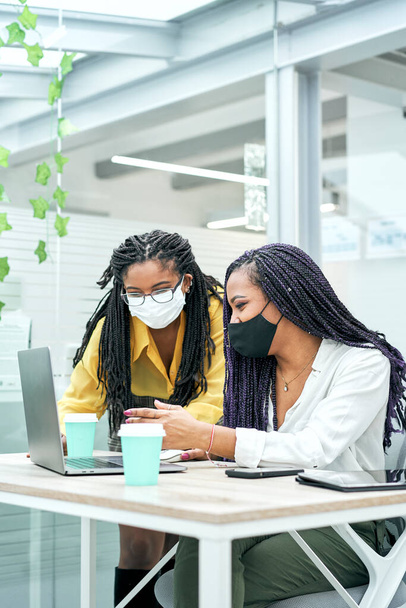 Függőleges lövés két mosolygós fiatal női vállalkozó visel arc maszk, miközben dolgozik a laptop az irodában. - Fotó, kép