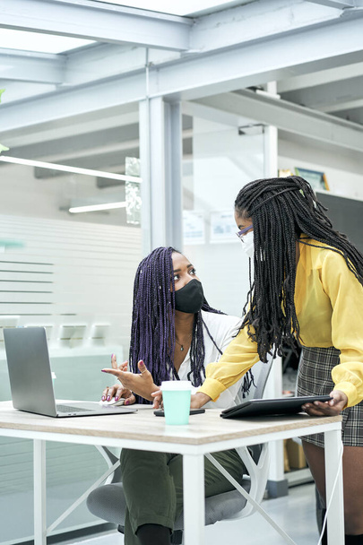 Plan vertical de deux jeunes entrepreneures portant un masque facial pendant qu'elles travaillent sur un ordinateur portable au bureau. - Photo, image