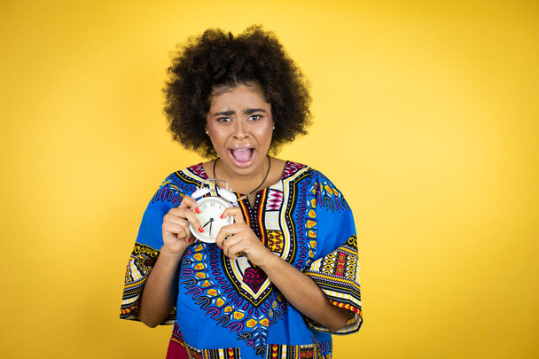 donna afro-americana indossa abiti africani su sfondo giallo urlando e spaventato, indicando l'orologio - Foto, immagini