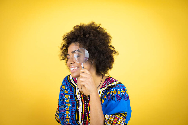 Afroamerykanka w afrykańskiej odzieży na żółtym tle zaskoczona patrząc przez lupę - Zdjęcie, obraz